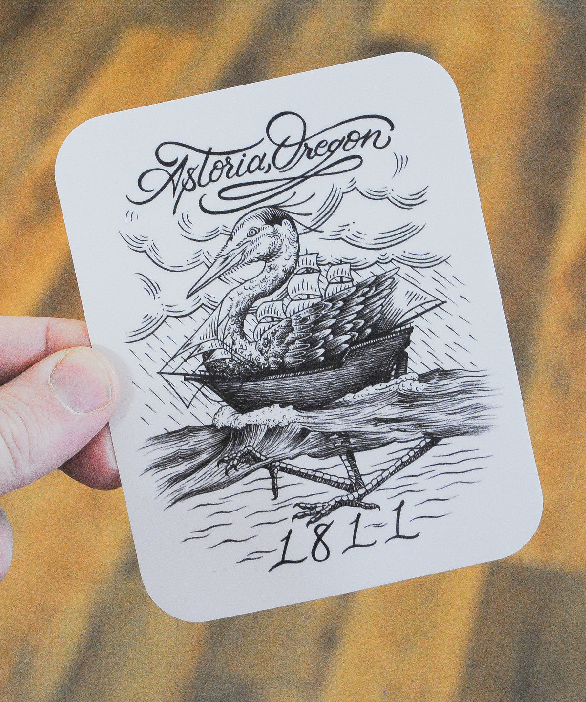 Black and white sticker with graphic of heron combined with a wooden ship on the water with rainstorm in the background in an etched styling, top of the sticker reads "Astoria, Oregon", bottom of the sticker reads "1811"