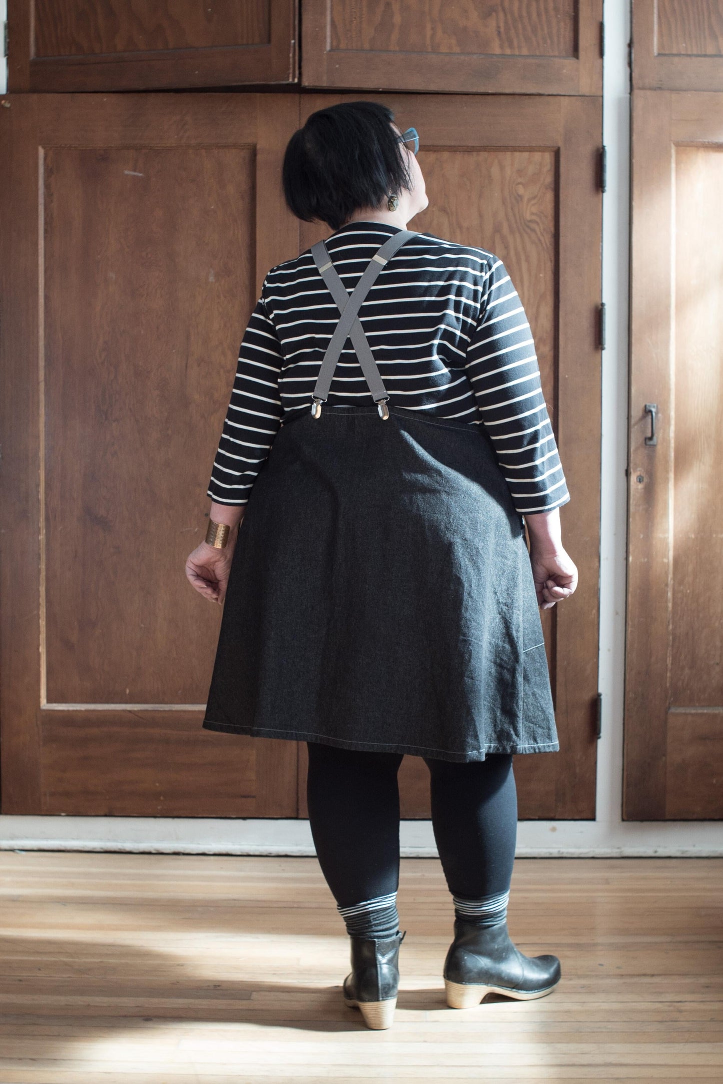 Back view of model wearing black denim suspender dress.100% cotton 8oz denim has a black wash and reads as very dark grey. The fabric is sturdy, hardworking yet lightweight. The pockets feature a contrasting self-trim.