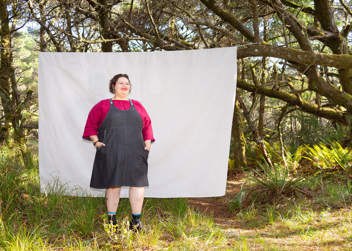 Model in forest with canvas backdrop wearing handmade black denim suspender dress with grey adjustable suspenders, knee-length apron pinafore style. Front view.
