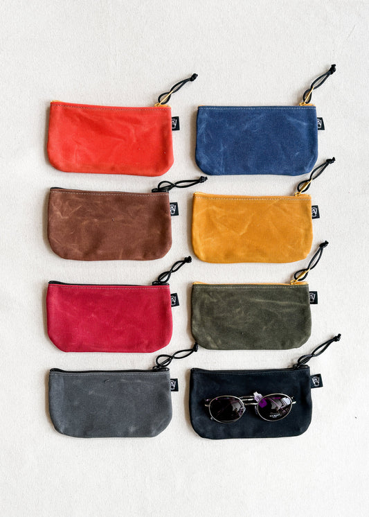8 assorted colorways of Shift + Wheeler handmade small waxed canvas zipper pouches, one with pair of sunglasses on top to detail size of pouches