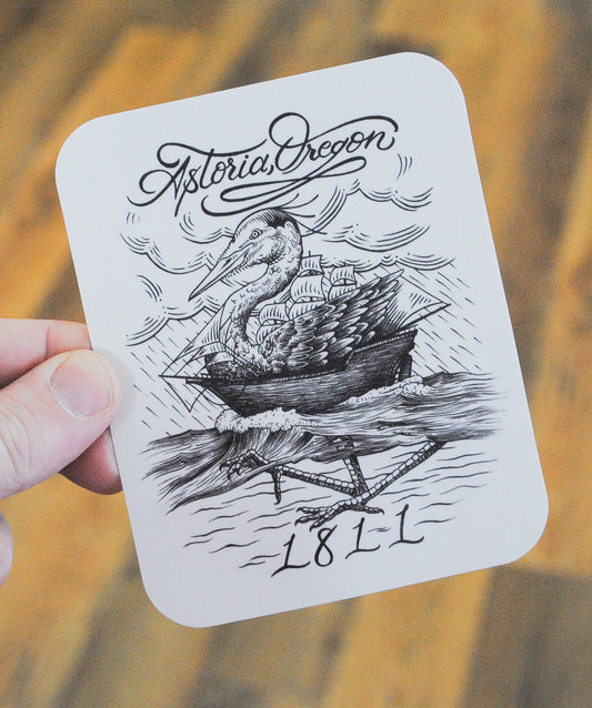 Black and white sticker with graphic of heron combined with a wooden ship on the water with rainstorm in the background in an etched styling, top of the sticker reads "Astoria, Oregon", bottom of the sticker reads "1811"