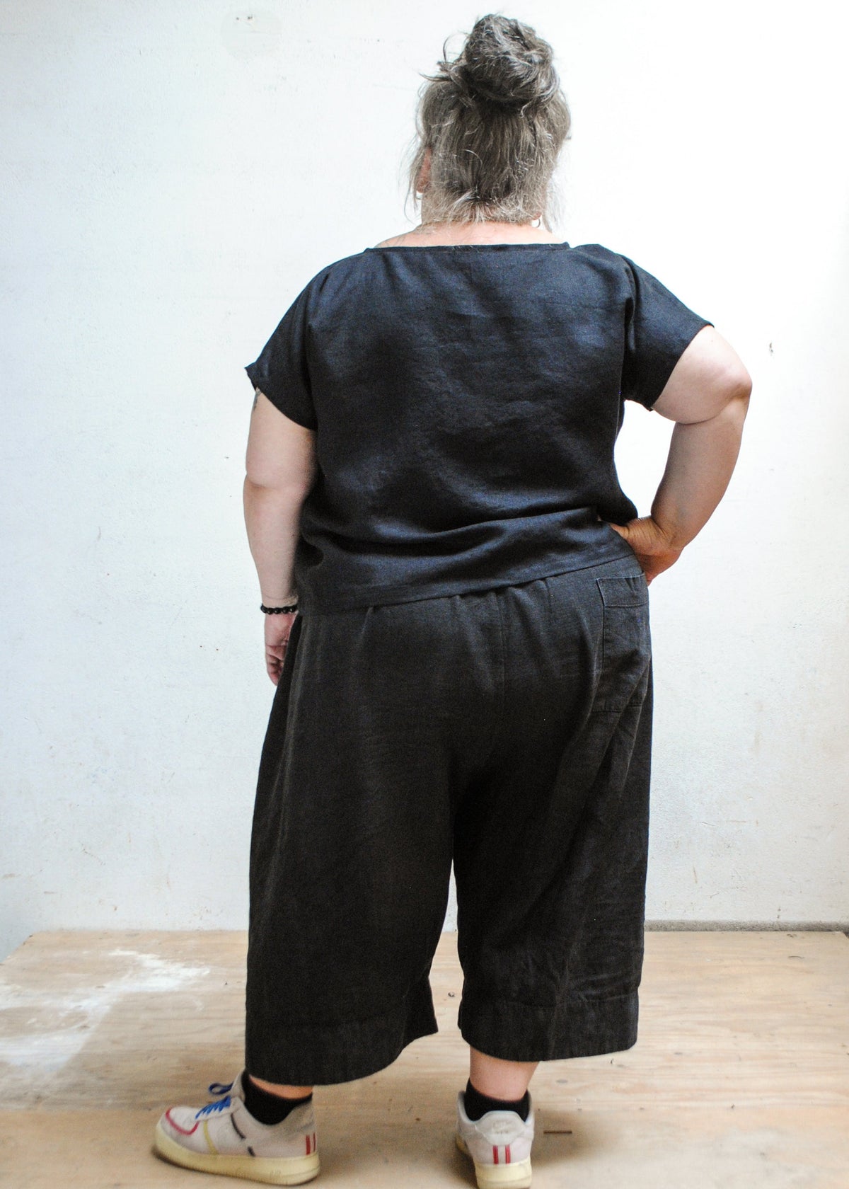 Model wearing Shift + Wheeler handmade black linen/cotton basic top with dark pants and tennis shoes, back view