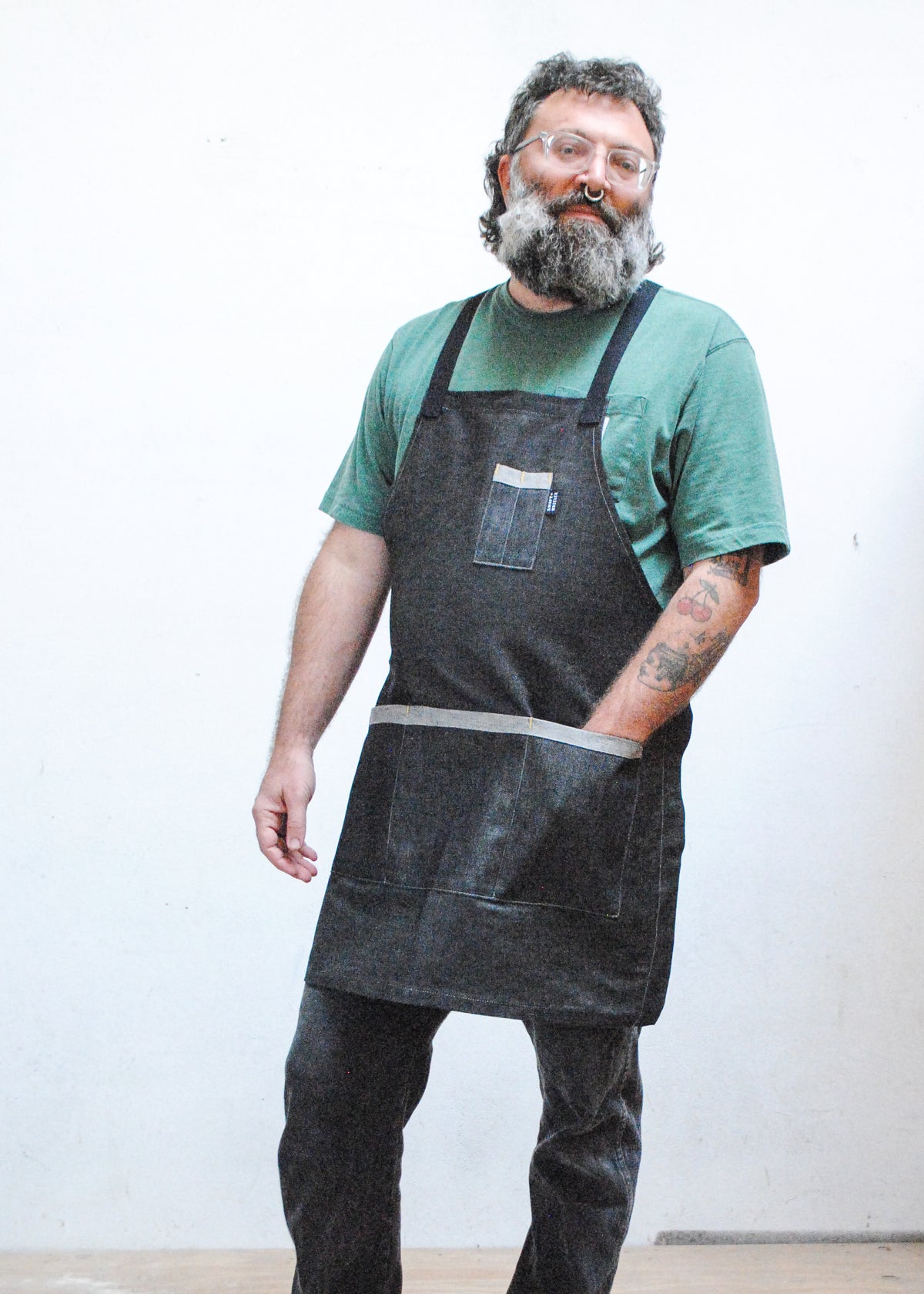 Model wearing sage green shirt, black denim pants, and black denim crossback apron, one hand in front apron pocket
