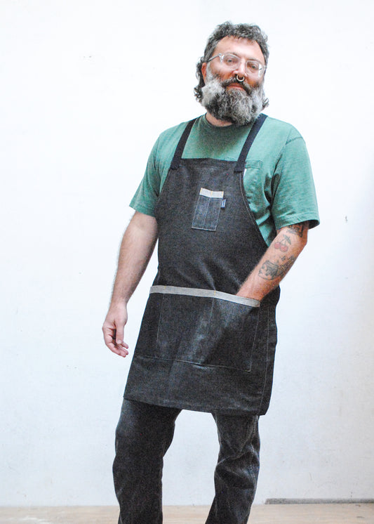 Model wearing sage green shirt, black denim pants, and black denim crossback apron, one hand in front apron pocket