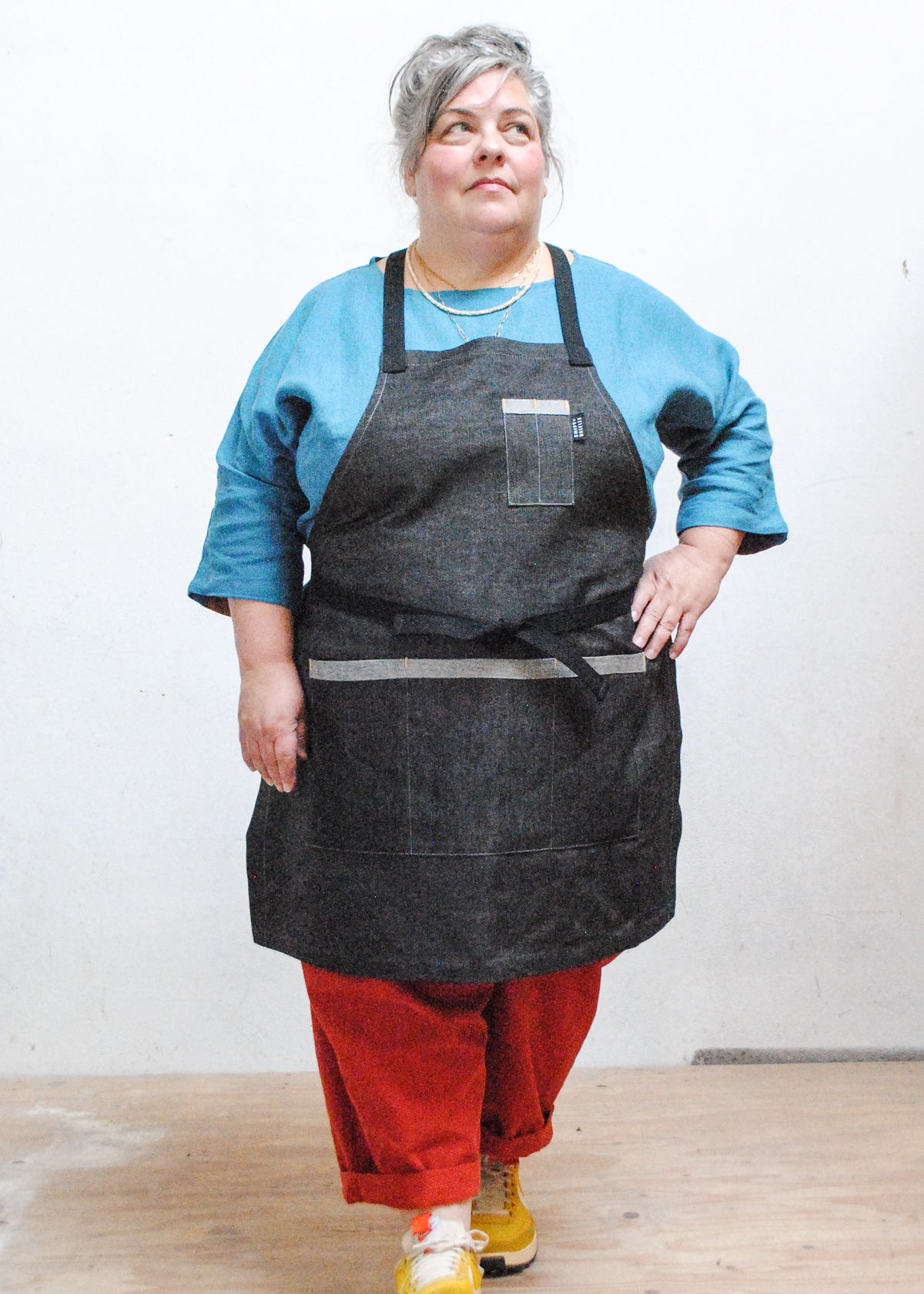 Model wearing marine blue shirt, red pants, sneakers, and black denim crossback apron, one hand on hip