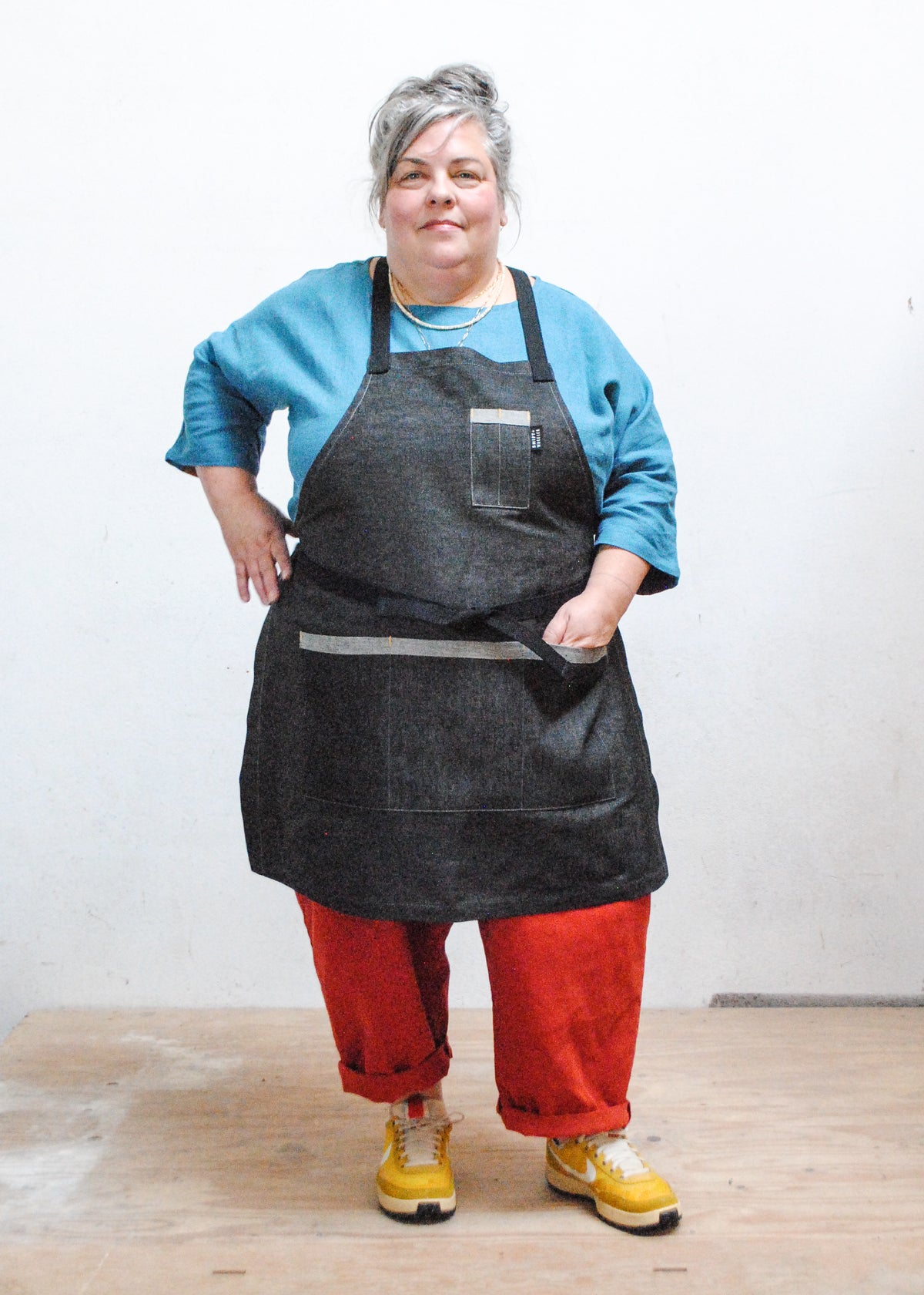 Model wearing marine blue shirt, red pants, sneakers, and black denim crossback apron