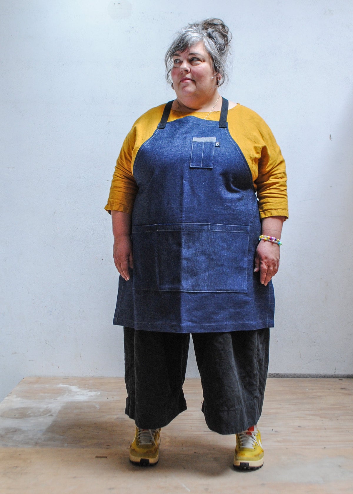 Model wearing yellow shirt, black pants, and Shift + Wheeler crossback denim apron, hands at sides