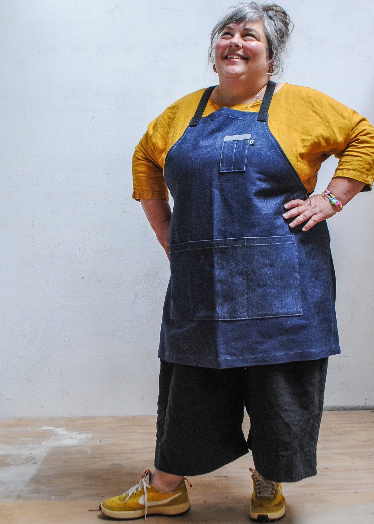 Model wearing yellow shirt, black pants, and Shift + Wheeler crossback denim apron, hands on hips