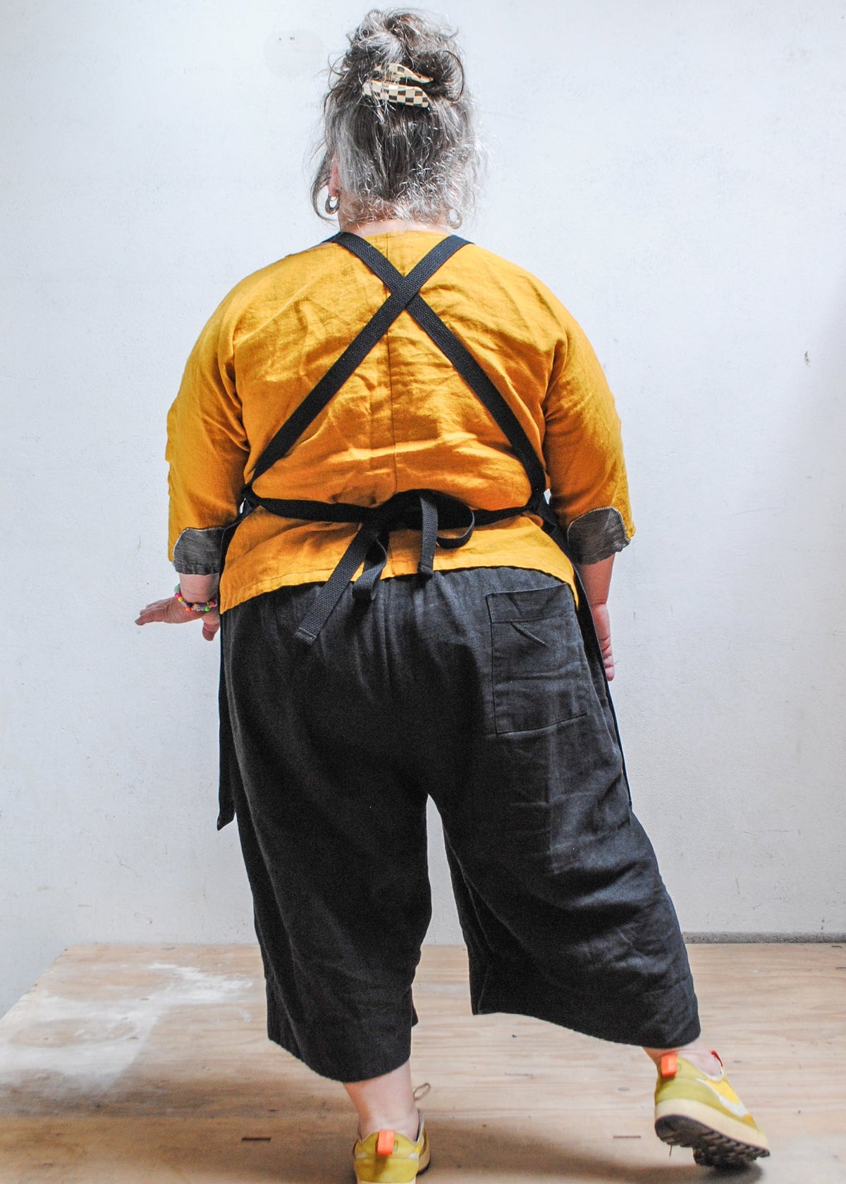 Model wearing yellow shirt, black pants, and Shift + Wheeler crossback denim apron, back view