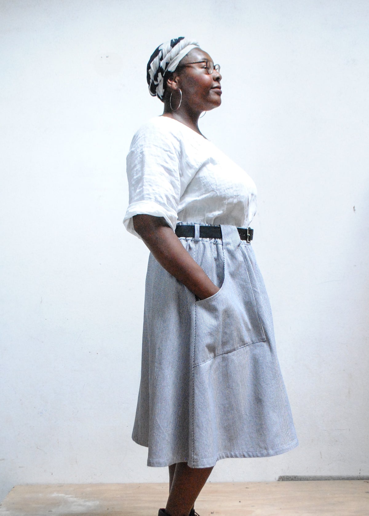 Model in white top, black boots, and Shift + Wheeler handmade railroad stripe skirt, hands in skirt pockets - side view