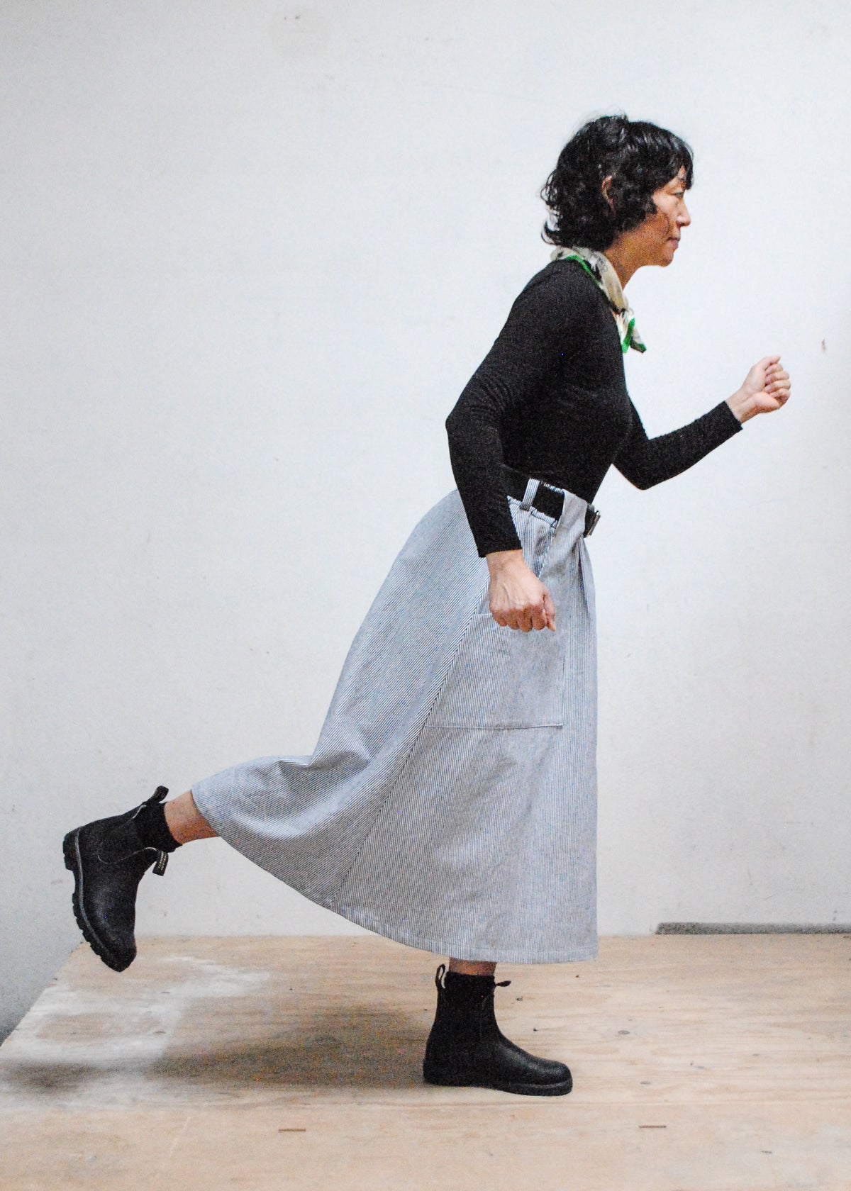 Model in black long sleeve shirt, black boots, and Shift + Wheeler handmade railroad stripe skirt, one foot off ground and arms in moving pose - side view