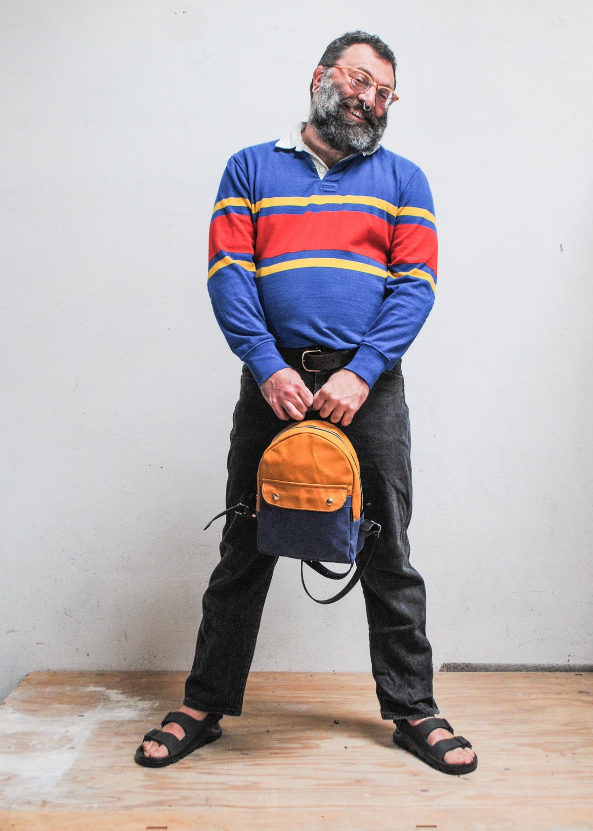 Model holding 2-tone yellow gold and royal handmade Shift + Wheeler waxed canvas mini backpack in front of them at arms length from backpack's top loop strap
