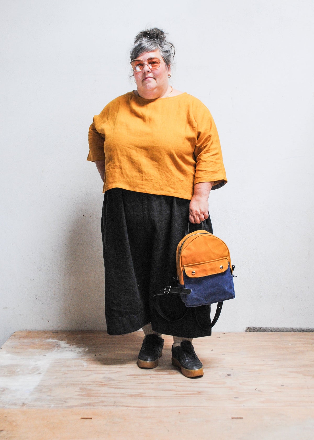 Model holding 2-tone yellow gold and royal handmade Shift + Wheeler waxed canvas mini backpack at arms length from backpack top strap