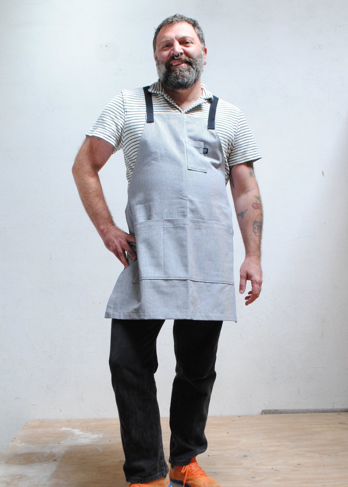Man with hand on hip wearing striped short sleeve collared shirt, dark pants, and handmade railroad stripe apron.