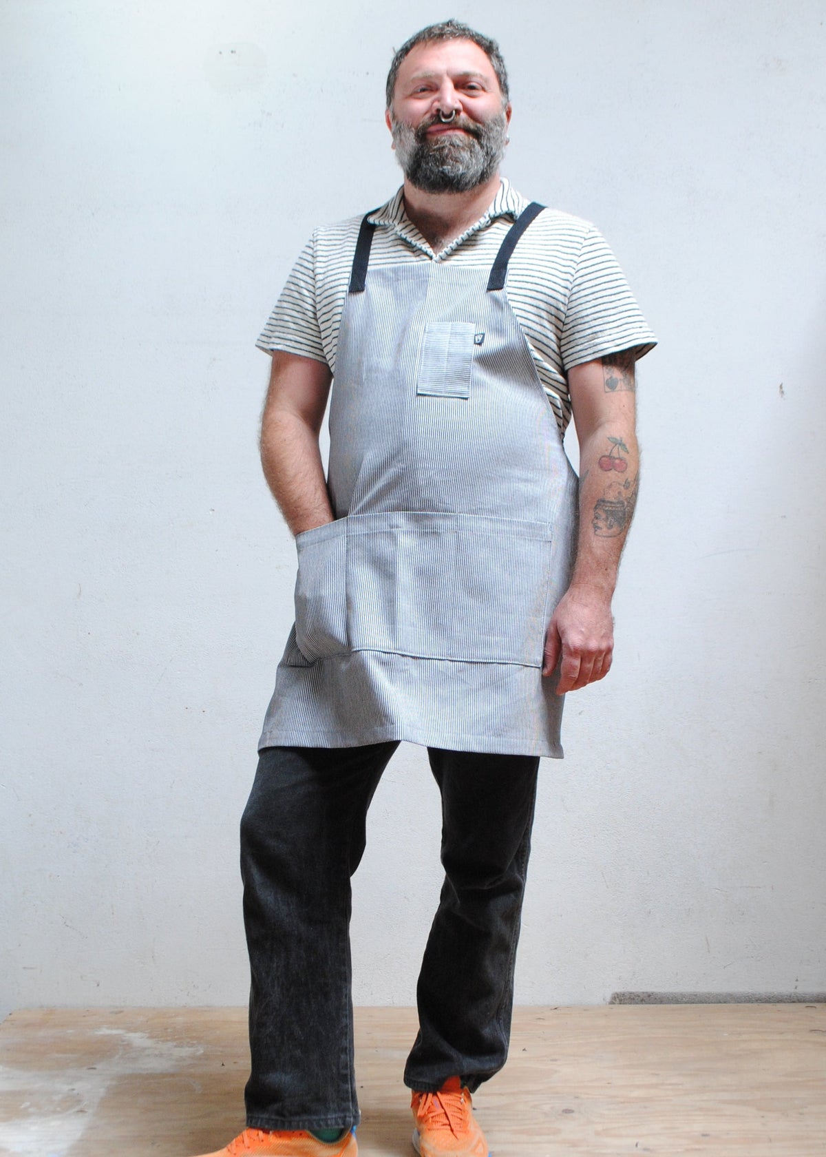 Man wearing striped short sleeve collared shirt, dark pants, and with hand in front pocket of handmade railroad stripe apron.