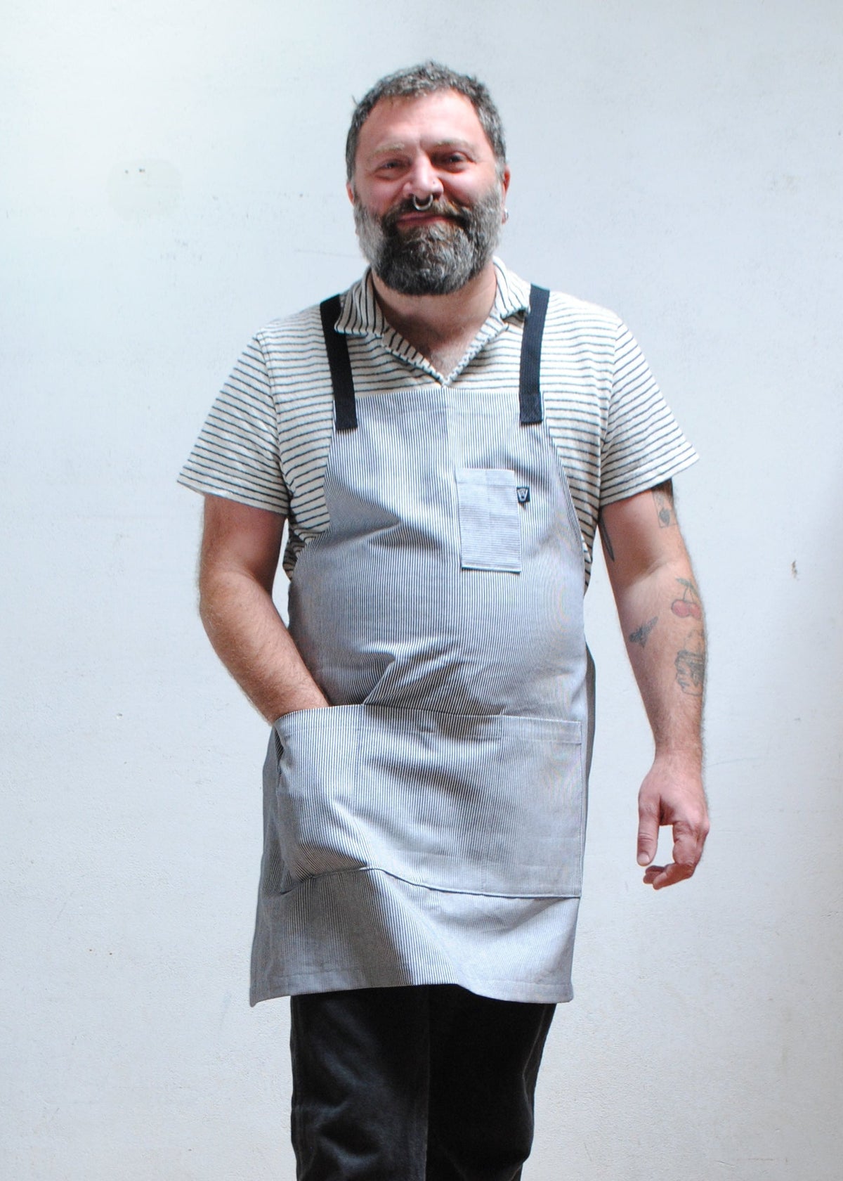 Man wearing striped short sleeve collared shirt, dark pants, with hand in front pocket of handmade railroad stripe apron.