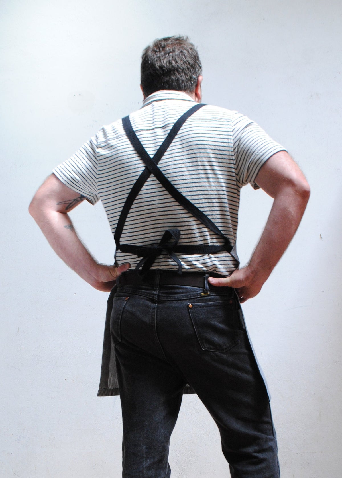 Man with hand on hips  wearing striped short sleeve collared shirt, dark pants, and handmade railroad stripe apron. Back view