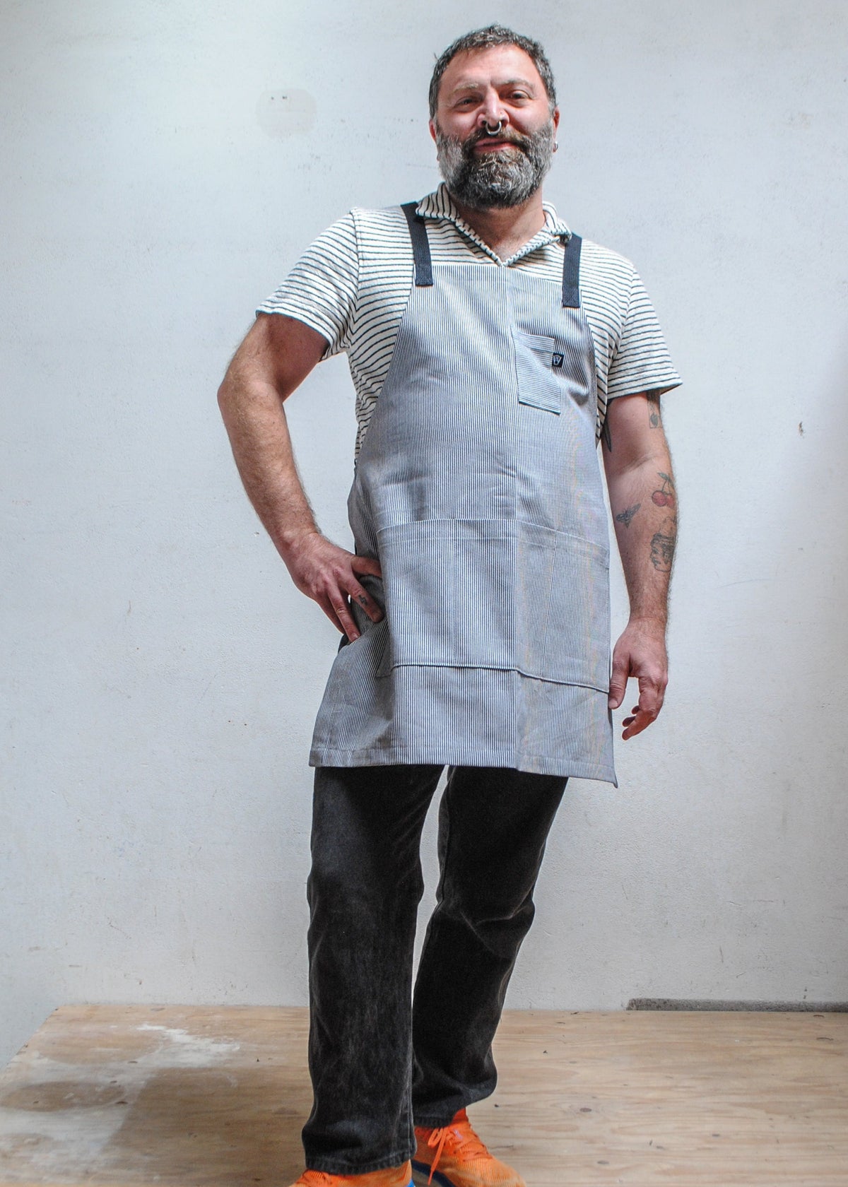 Man with hand on hip wearing striped short sleeve collared shirt, dark pants, and handmade railroad stripe apron.
