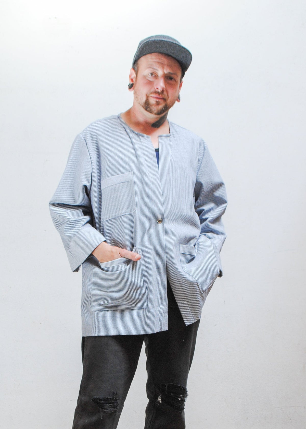 Model wearing a gray hat, dark pants, and railroad stripe button-up lab coat style jacket with hands in front pockets