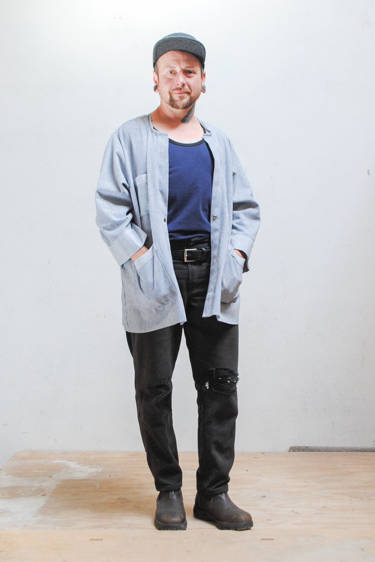 Model wearing a gray hat, dark pants, and railroad stripe button-up lab coat style jacket with hands in front pockets. Full length view with jacket unbuttoned.