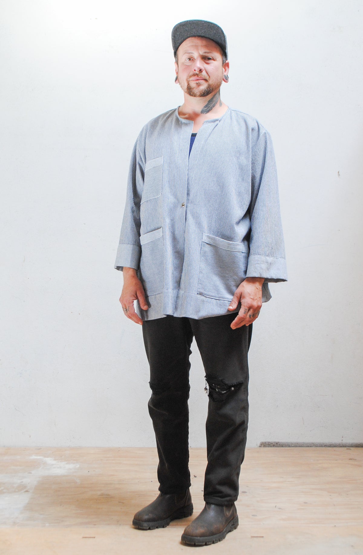 Model wearing a gray hat, dark pants, and railroad stripe button-up lab coat style jacket. Full length view with jacket buttoned