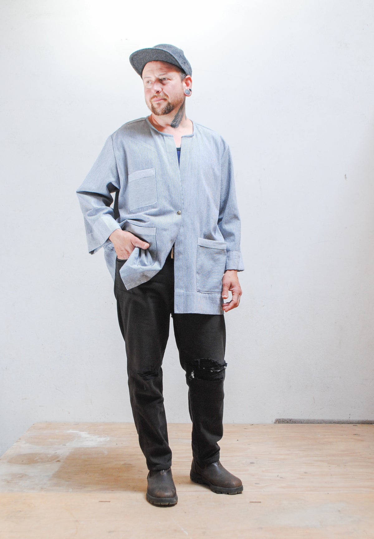 Model wearing a gray hat, dark pants, and railroad stripe button-up lab coat style jacket with one hand in front pant pocket.