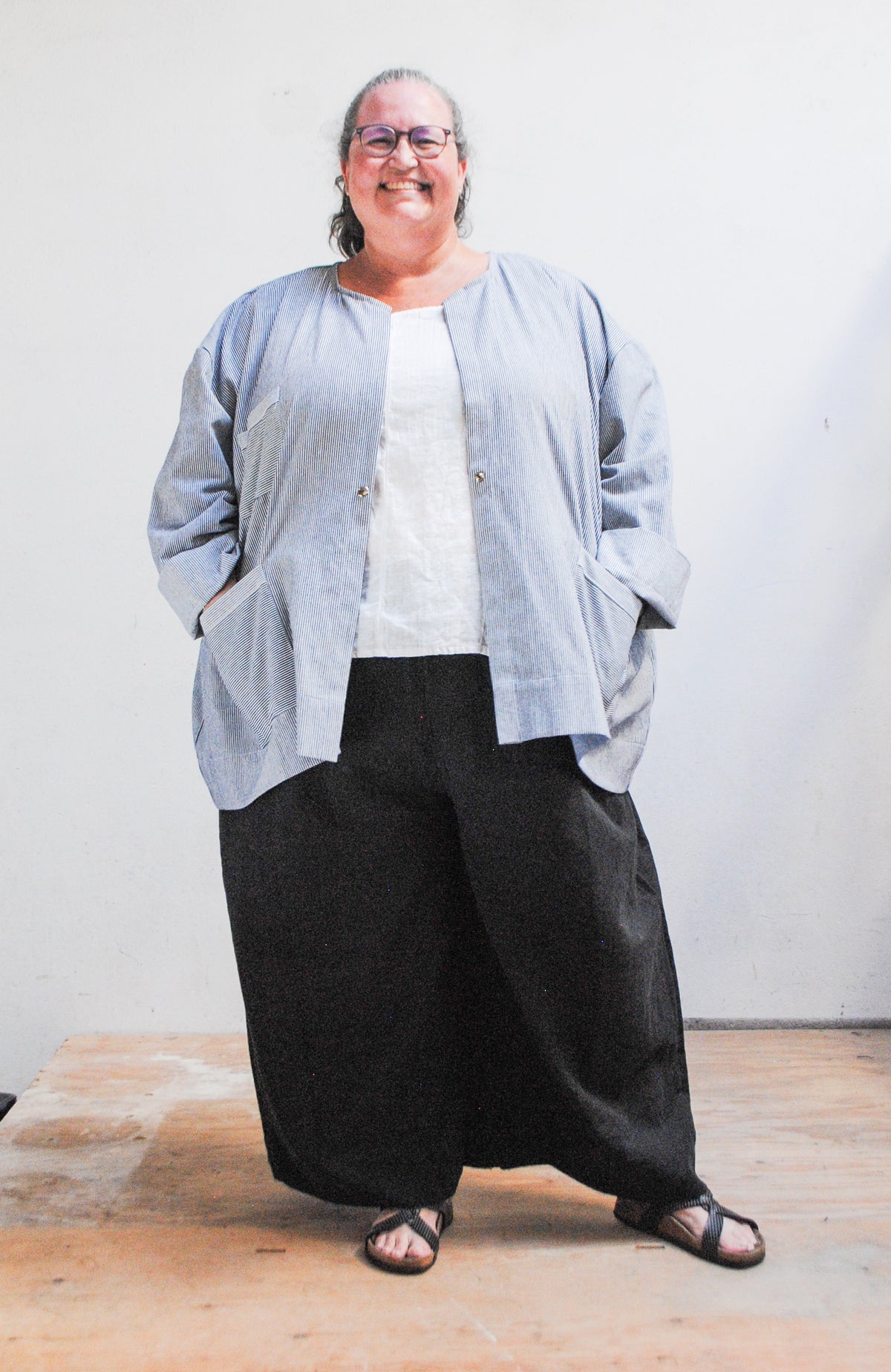 Model wearing a gray hat, dark pants, and railroad stripe button-up lab coat style jacket with hands in front pockets of jacket. Front view with jacket unbuttoned.