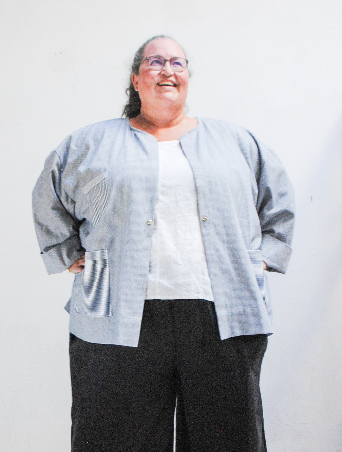 Model wearing a gray hat, dark pants, and railroad stripe button-up lab coat style jacket with hands on hips. Front view with jacket unbuttoned.