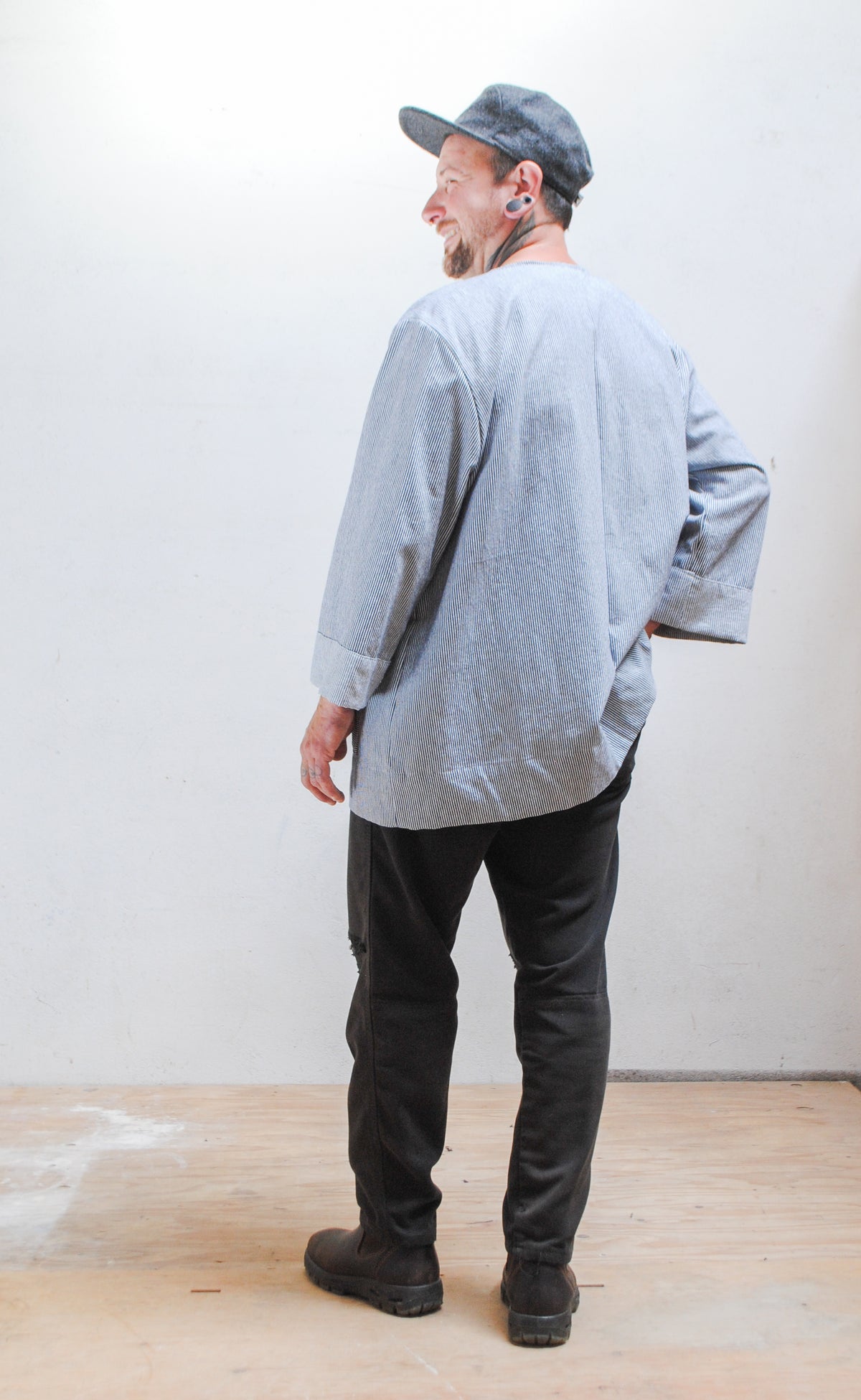 Model wearing a gray hat, dark pants, and railroad stripe button-up lab coat style jacket with hands in front pant pocket. Back view.
