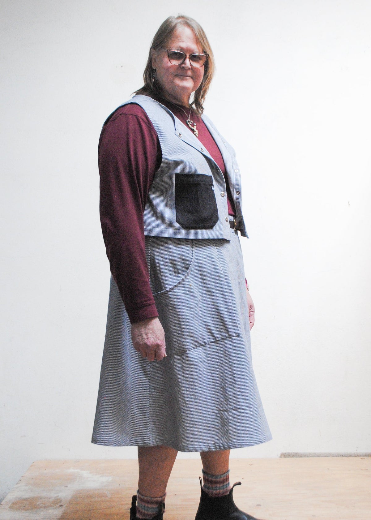 Model in burgundy long sleeve shirt, railroad strip vest, boots, and Shift + Wheeler handmade railroad stripe skirt - side view