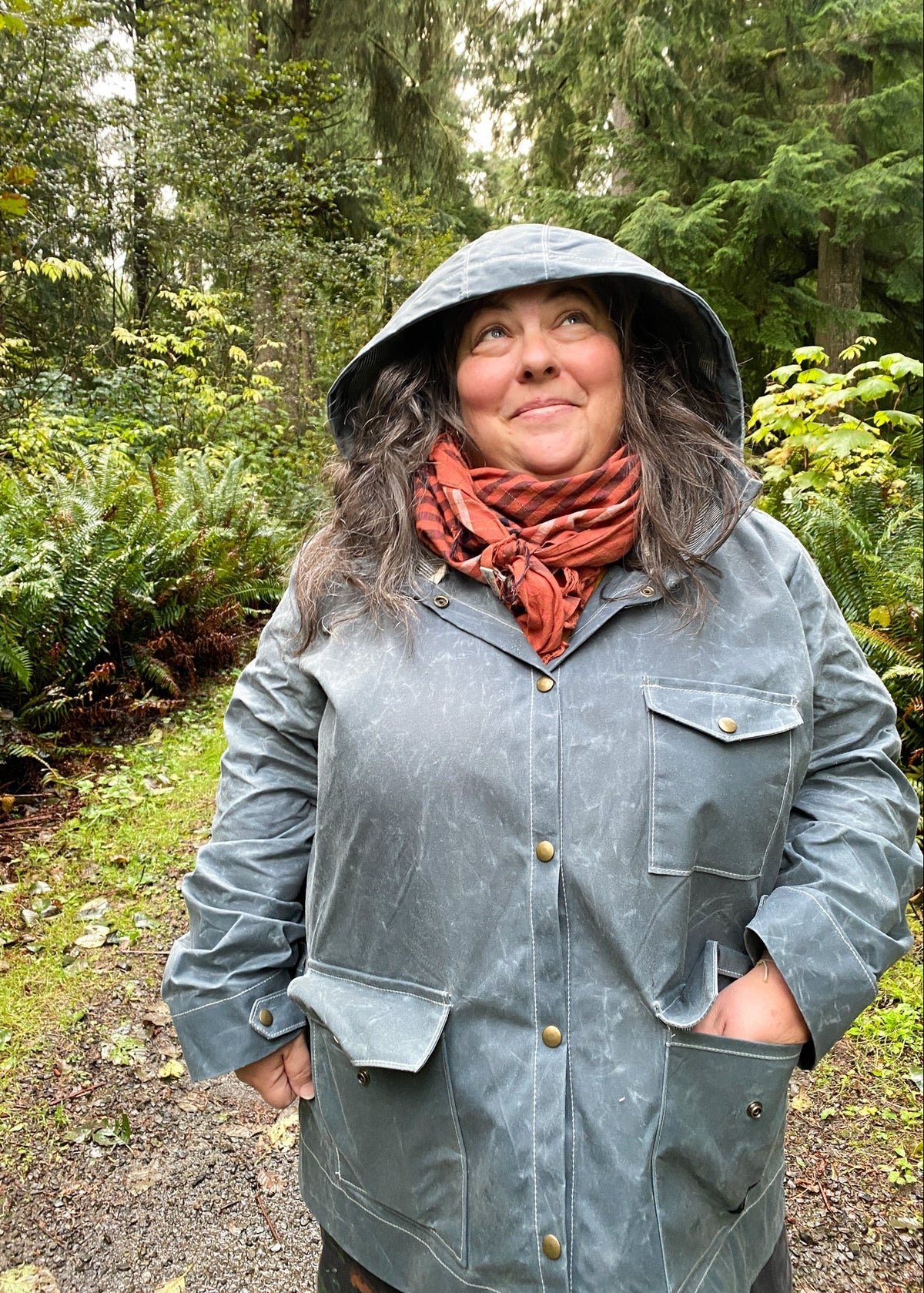 A woman wearing slate waxed canvas hooded rain jacket with hood on and hand in one pocket of jacket, standing outdoors in lush forest.