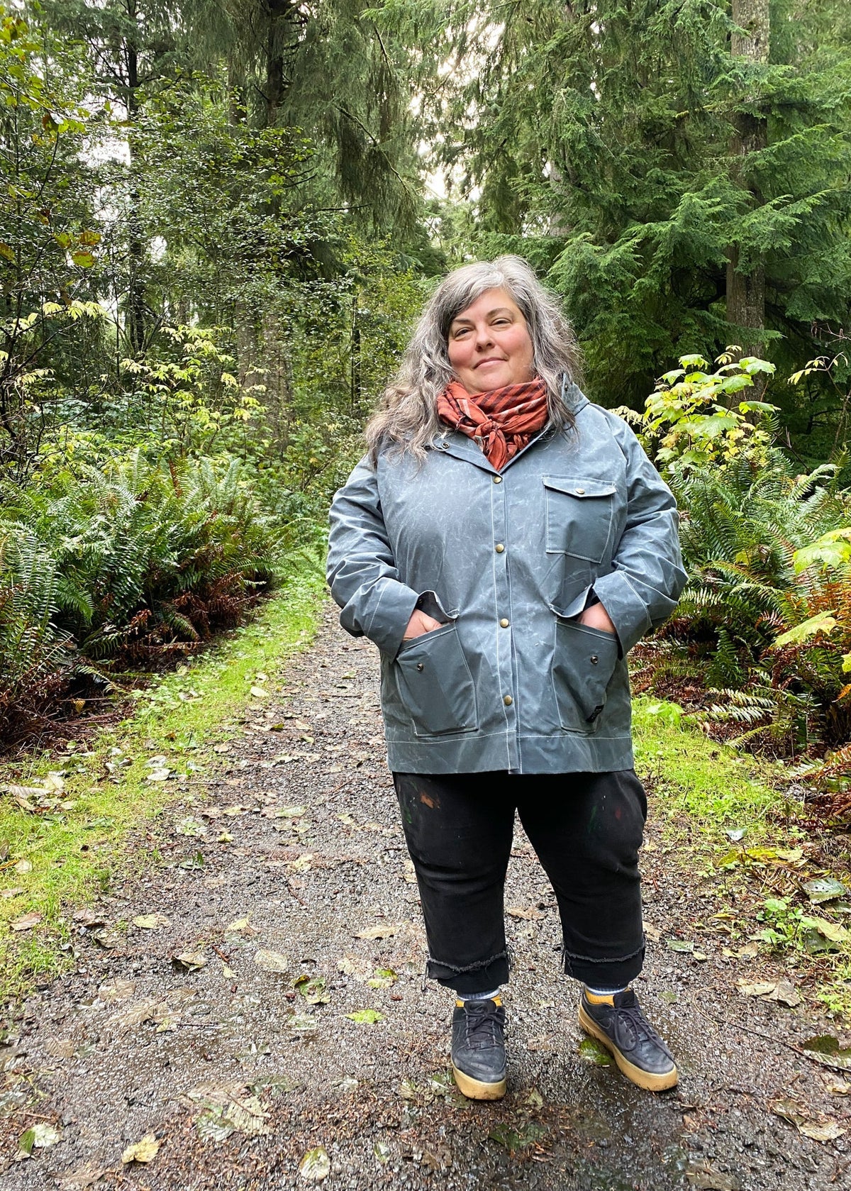 A woman standing on a gravel path in a lush green forest, wearing a slate handmade waxed canvas rain jacket, a rust-orange scarf, black pants, and gray sneakers with yellow soles.