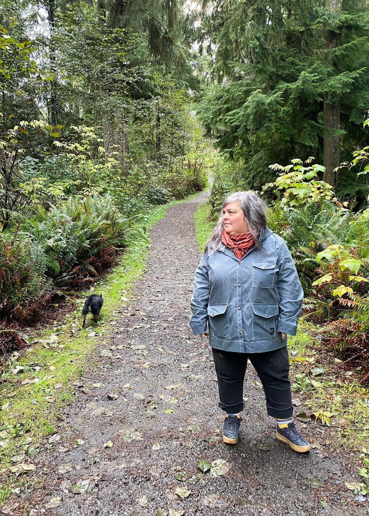 A woman standing on a gravel path wearing a slate handmade waxed canvas rain jacket, dark pants, and a red scarf, with a small black dog walking beside her through a lush green forest setting.
