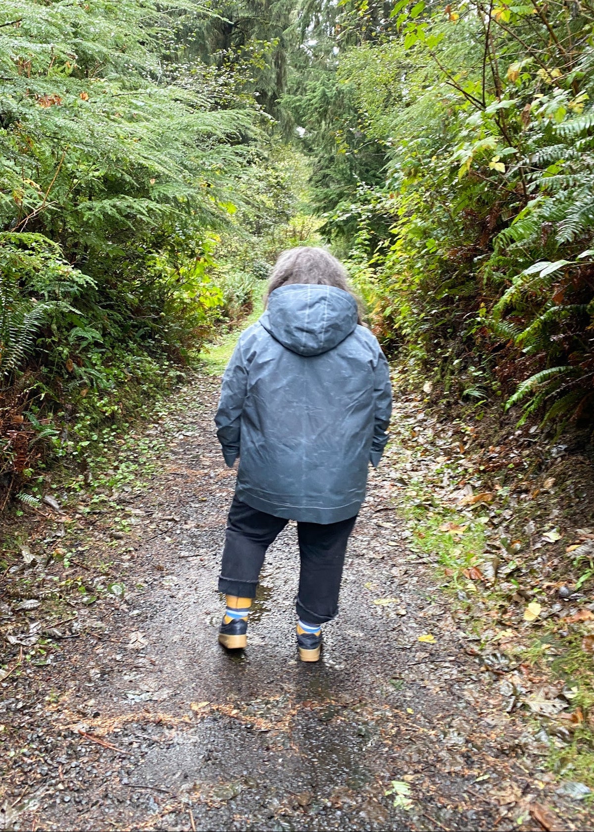 A woman standing on a forest path wearing a slate handmade waxed canvas rain jacket and dark pants. Back view.