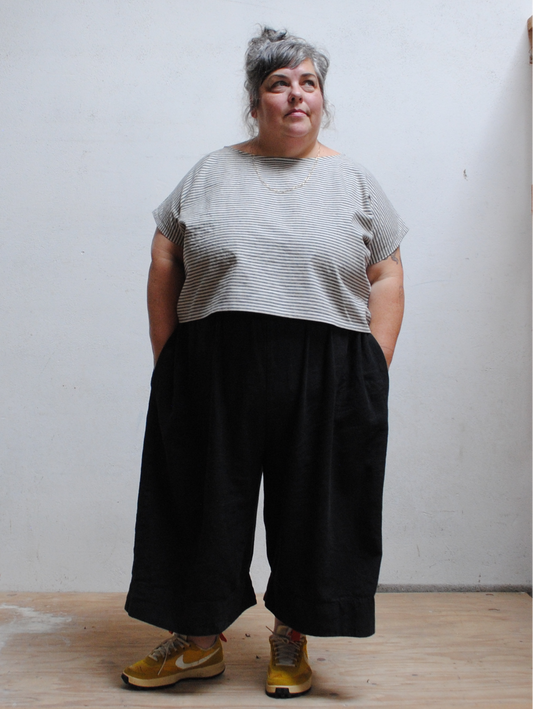 Person wearing a gray striped shirt and black wide-leg pants against a white wall.