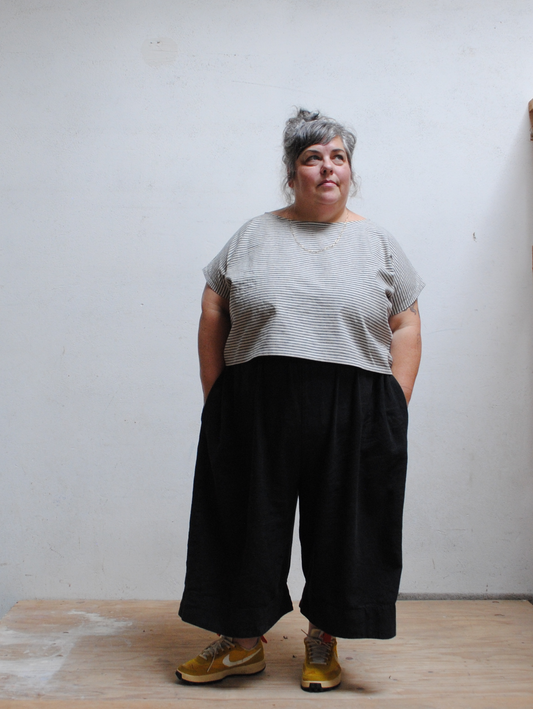 Person wearing a gray striped shirt and black wide-leg pants against a white wall.