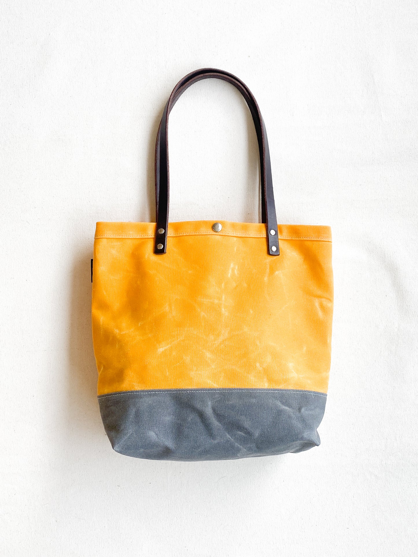 A waxed canvas utility tote with bridle leather handles, featuring a yellow upper half and gray lower half, with two side pockets.