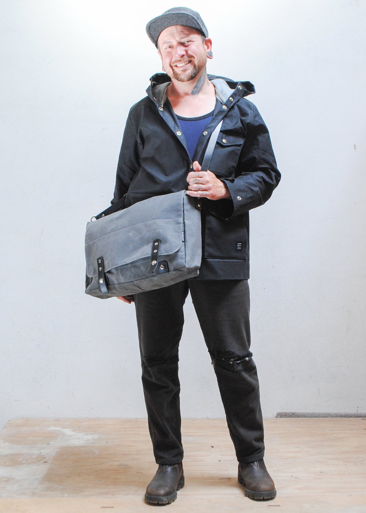 Model wearing black Shift + Wheeler black raincoat and charcoal Shift + Wheeler waxed canvas messenger bag over the front of shoulder
