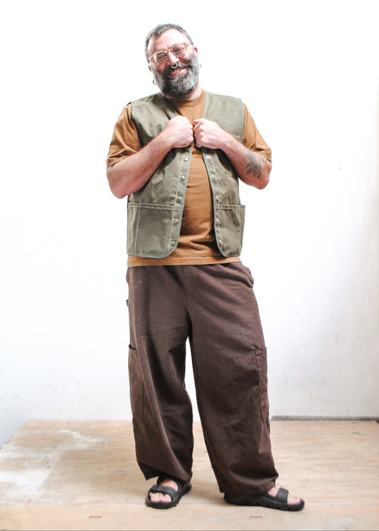 A man with a beard and glasses wearing a brown shirt, an olive green waxed canvas vest, loose brown pants, and black sandals.
