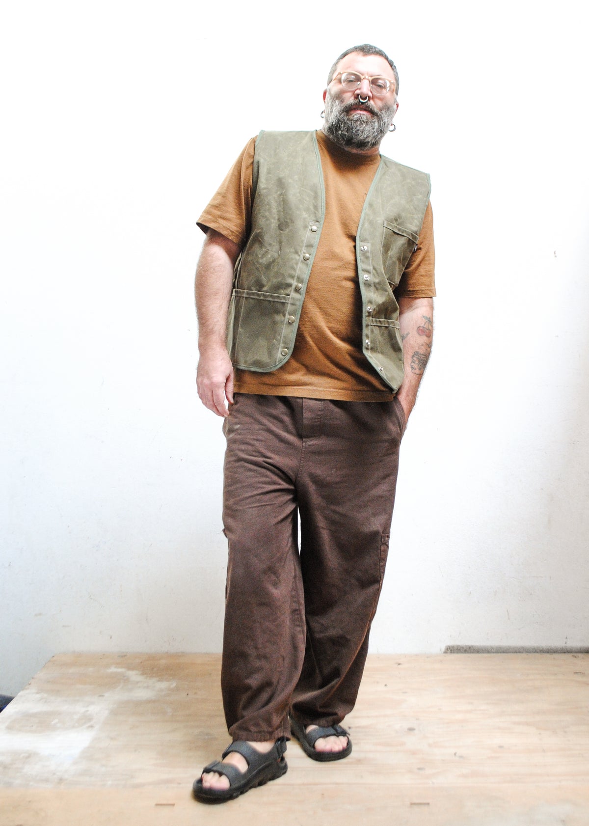 A man with a beard and glasses wearing a brown shirt, an olive green waxed canvas vest, loose brown pants, and black sandals.