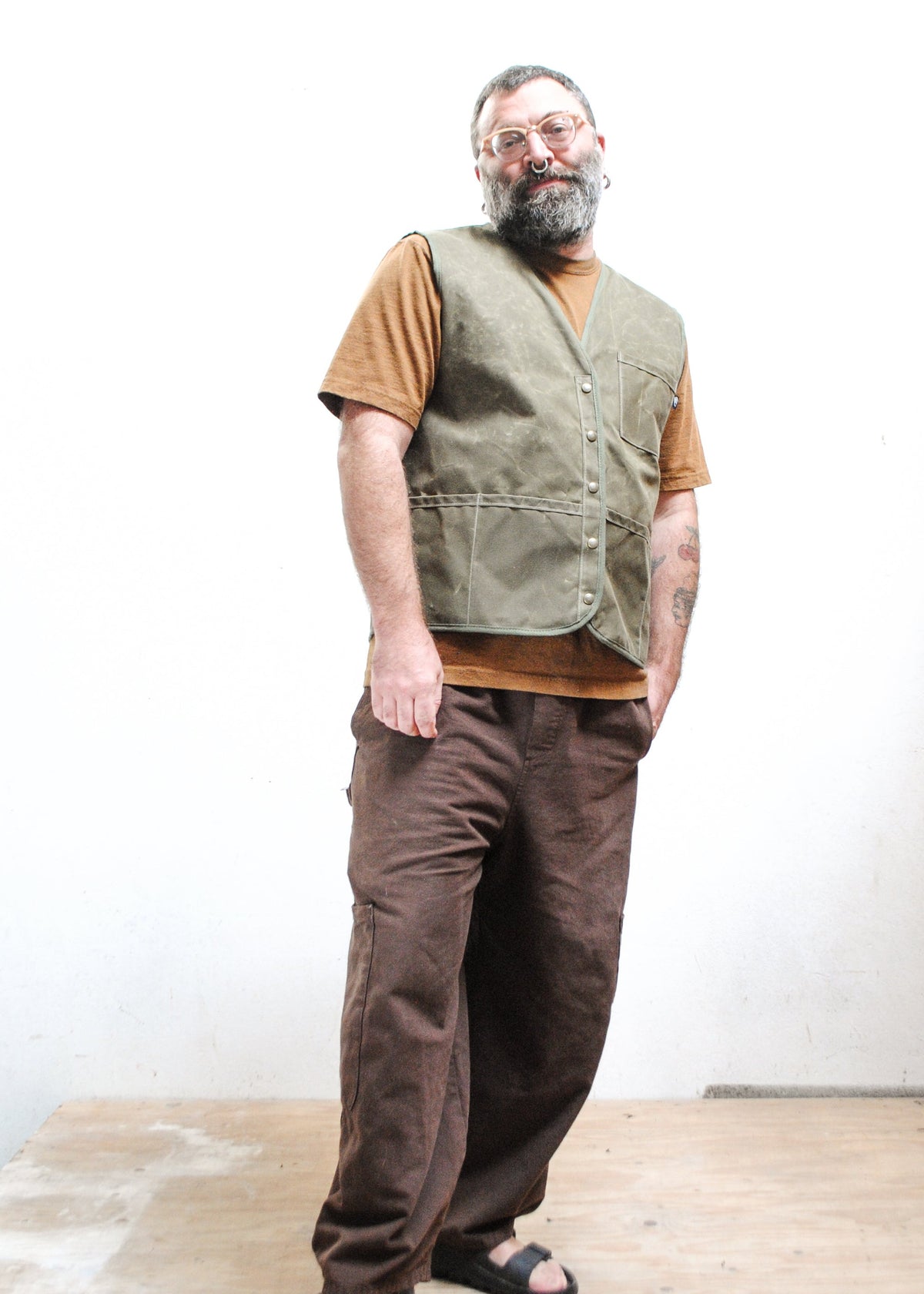A man with a beard and glasses wearing a brown shirt, an olive green waxed canvas vest, loose brown pants, and black sandals.