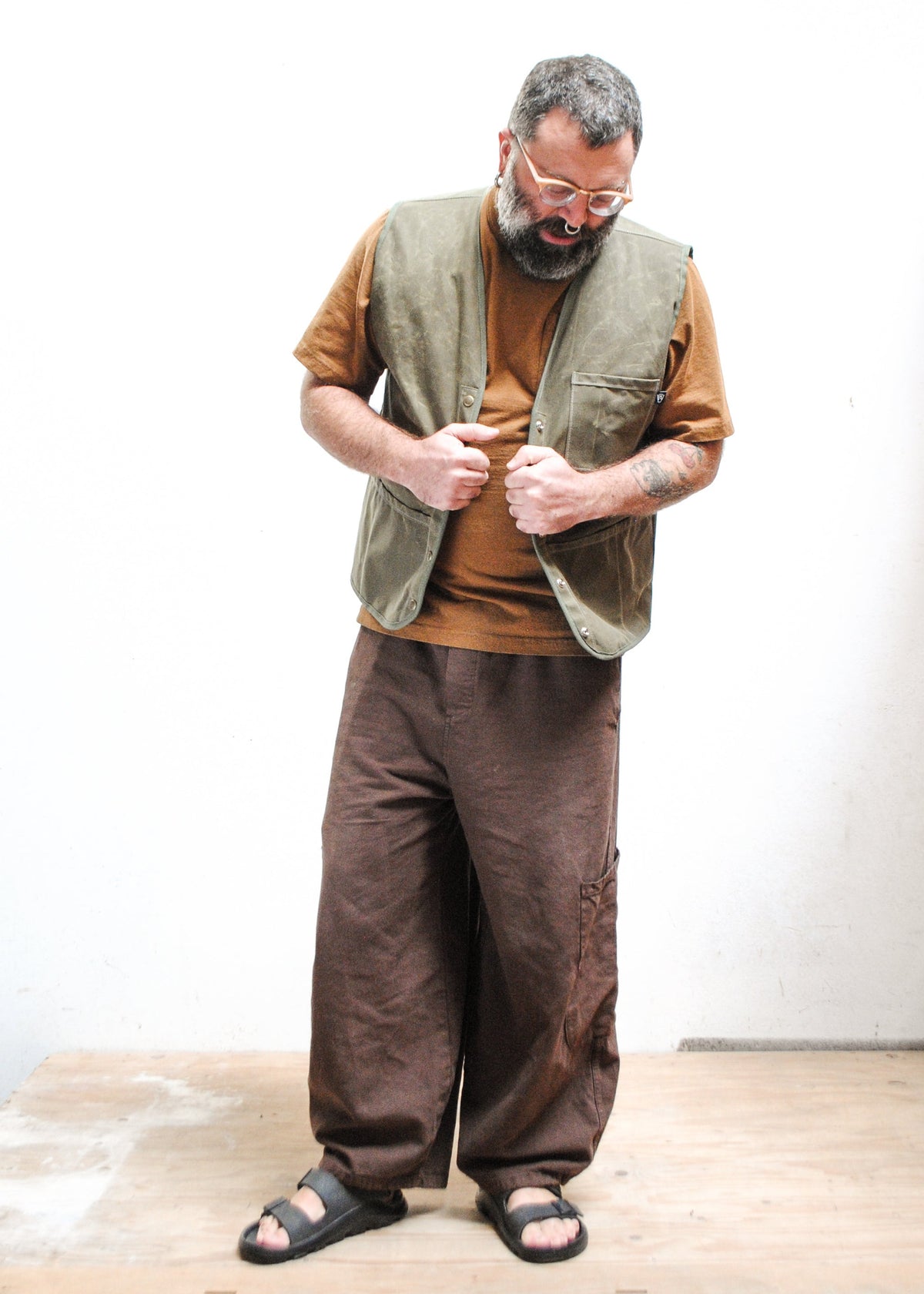 A man with a beard and glasses wearing a brown shirt, an olive green waxed canvas vest, loose brown pants, and black sandals.