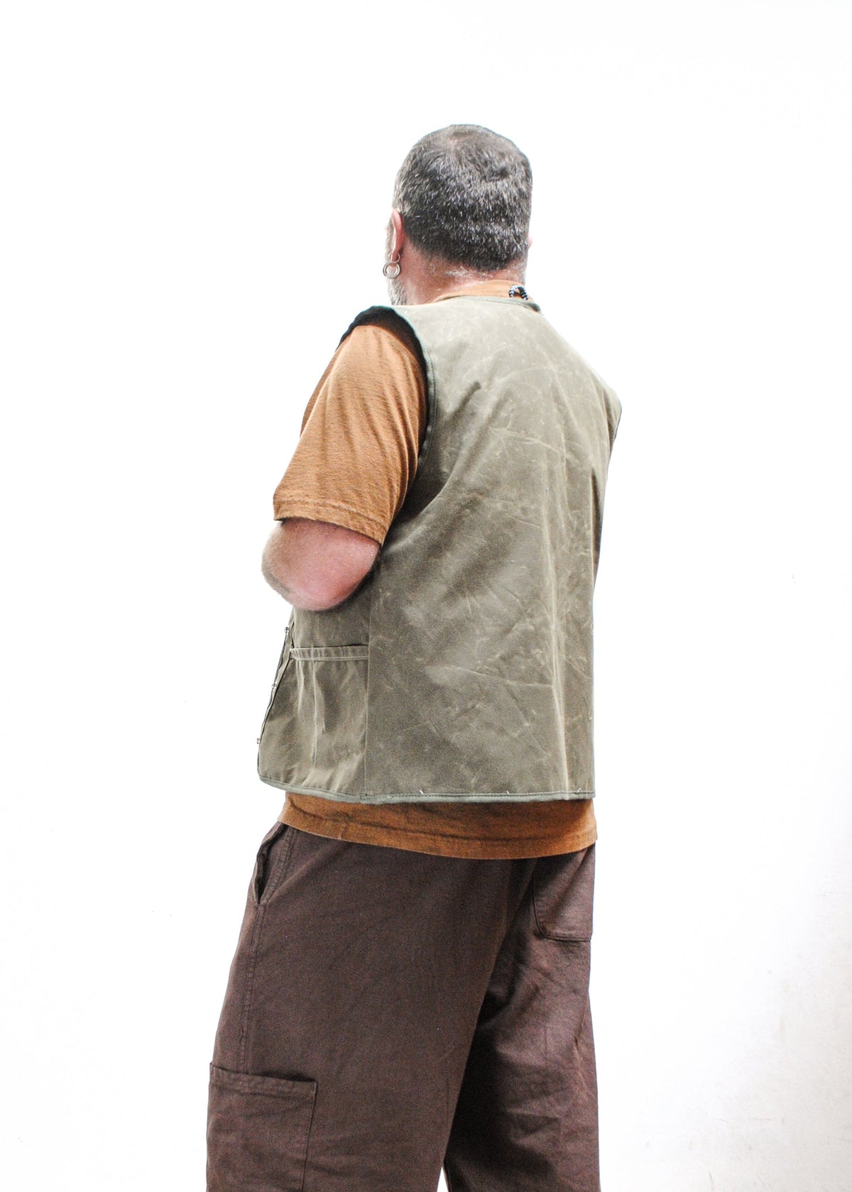 A man with a beard and glasses wearing a brown shirt, an olive green waxed canvas vest. Back view