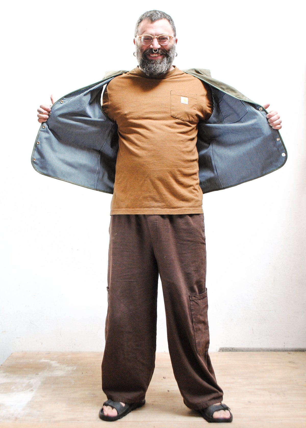 A man with a beard and glasses wearing a brown shirt, open view of inner railroad stripe lining of olive green waxed canvas vest, loose brown pants, and black sandals. 