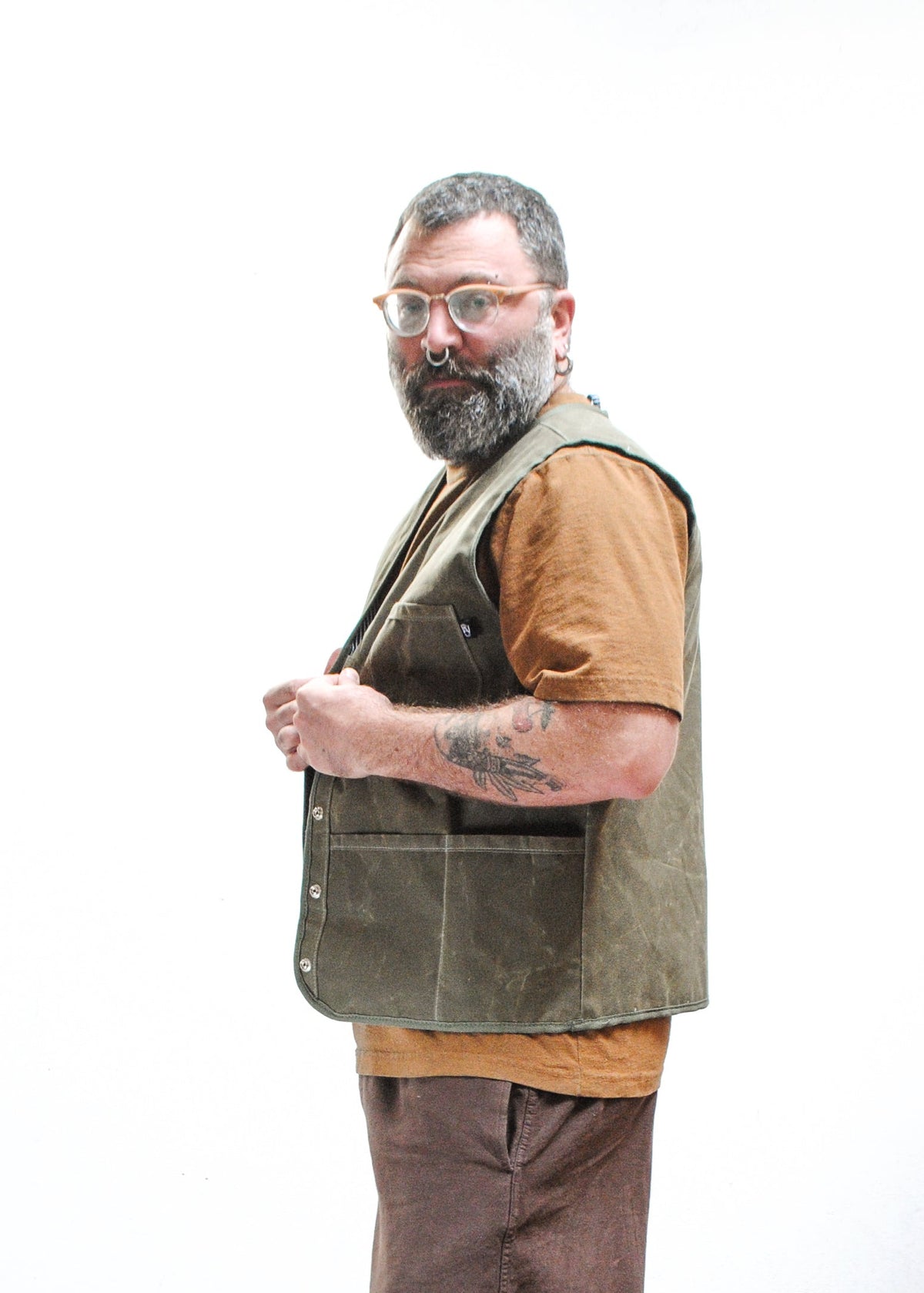 A man with a beard and glasses wearing a brown shirt and olive green waxed canvas vest. Side view.