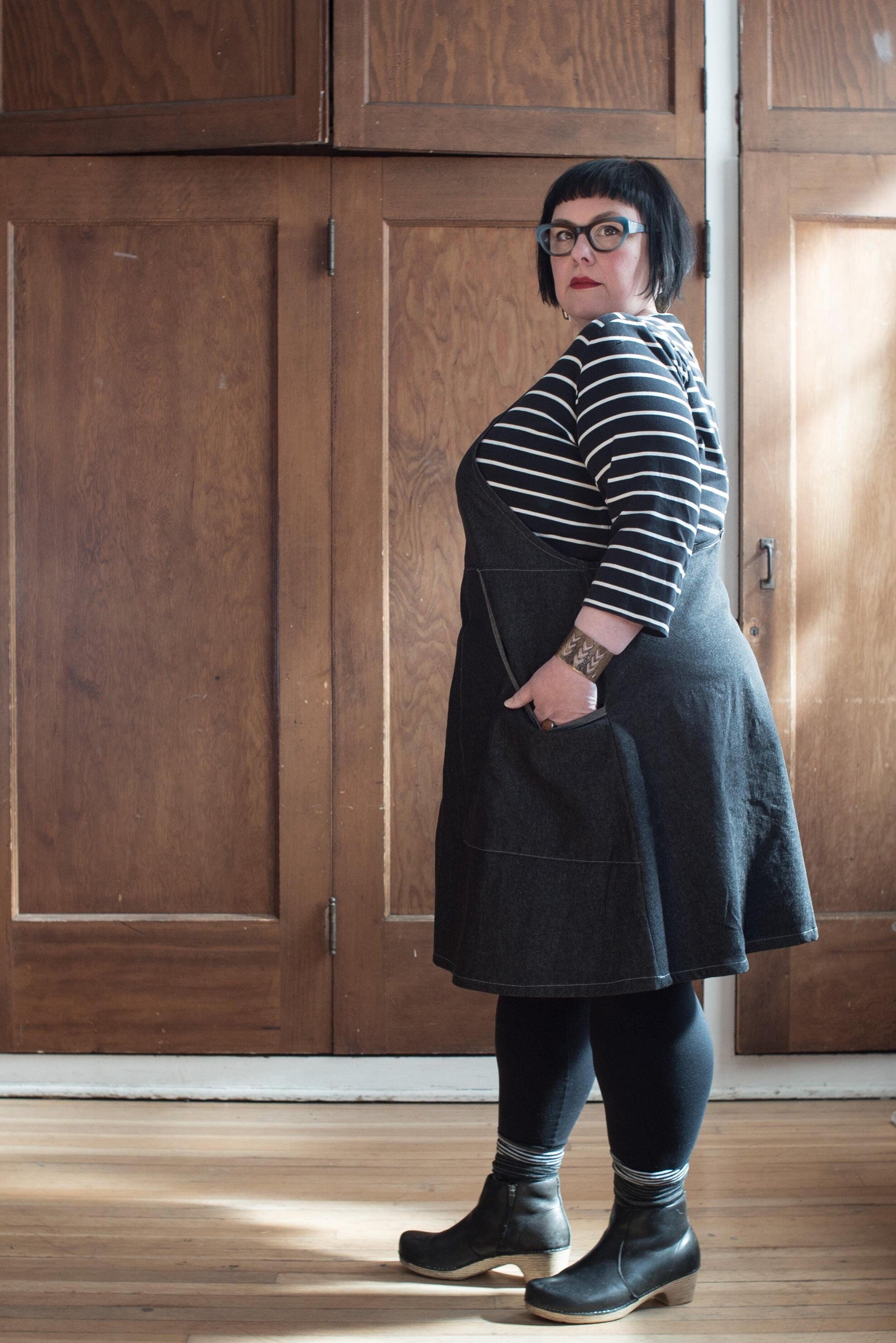 Side view of model wearing black denim suspender dress. 100% cotton 8oz denim has a black wash and reads as very dark grey. The fabric is sturdy, hardworking yet lightweight. The pockets feature a contrasting self-trim. 