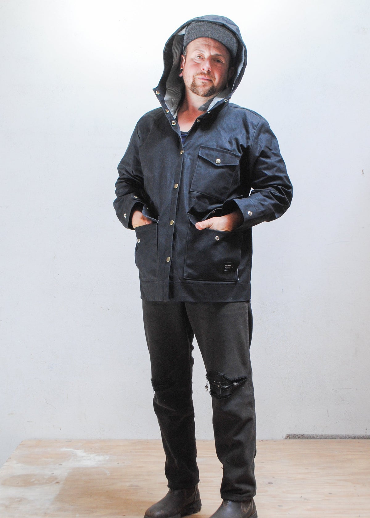 Model wearing black waxed canvas rain jacket with hood up and hands in front pockets