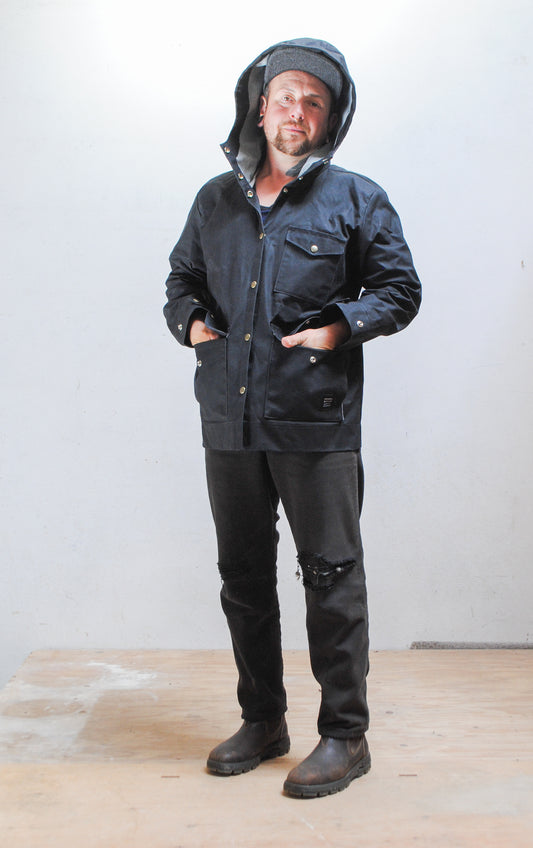 Model wearing black waxed canvas rain jacket with hood up and hands in front pockets
