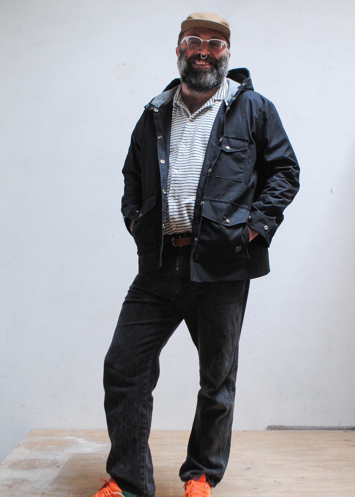 Model wearing black waxed canvas rain jacket, unbuttoned with hands tucked into waist pockets