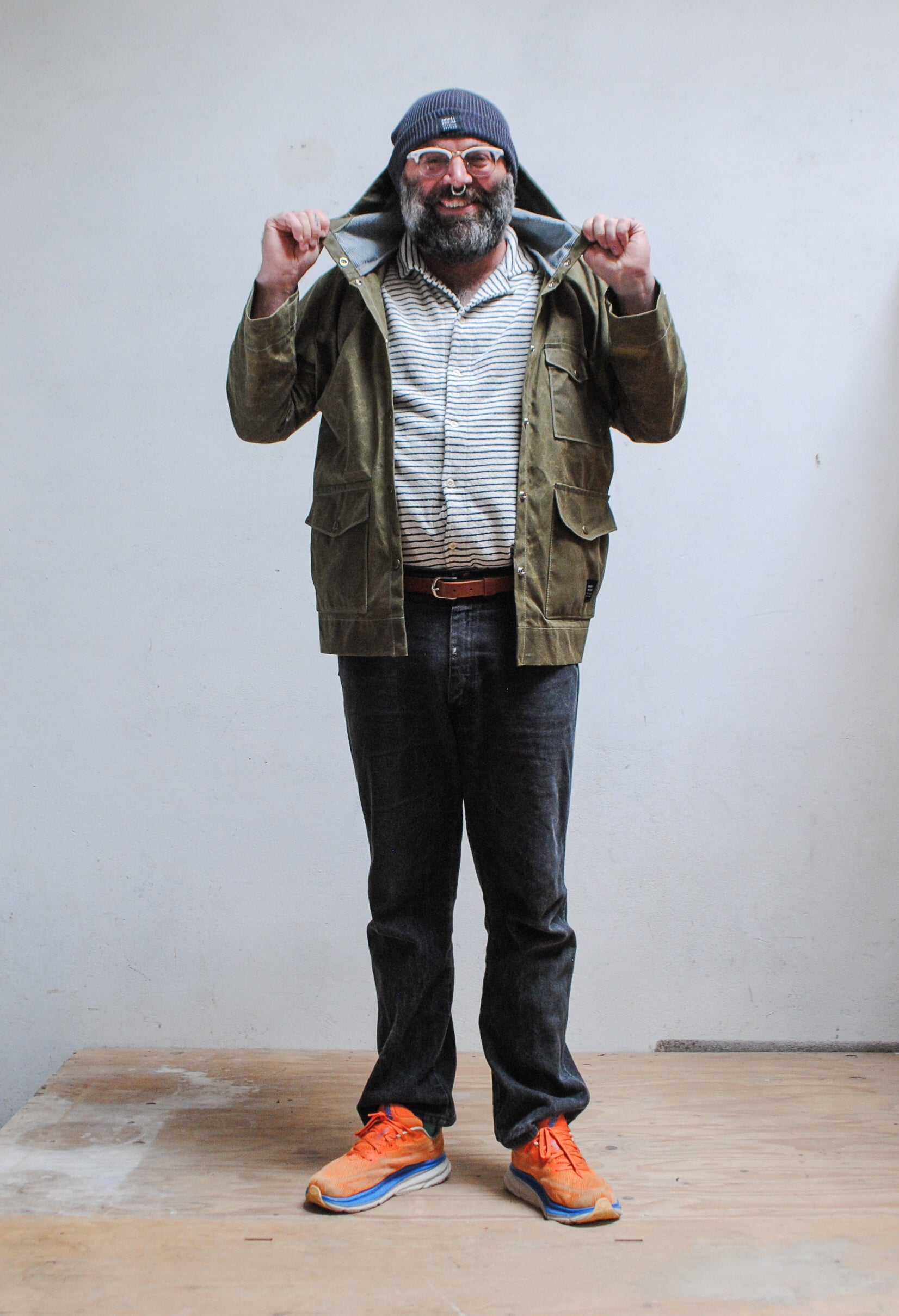 Man wearing a green waxed canvas jacket, striped shirt, jeans, and orange shoes against a plain background